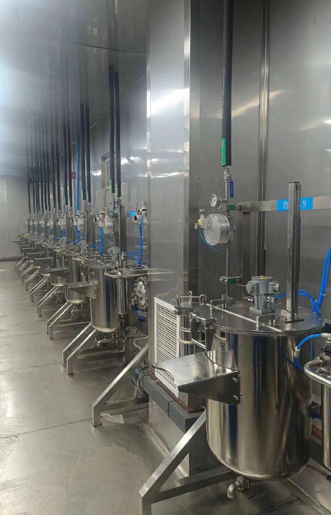 Row of stainless steel paint circulation tanks along the wall of a paint mixing room, each with its own colour station