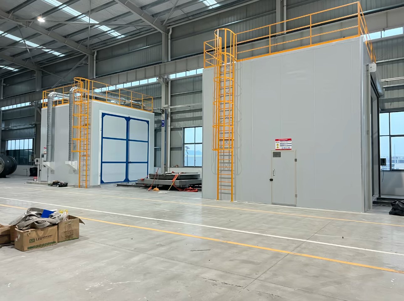 Front view of a sandblasting room with blue-framed doors and exhaust ducts beside a larger blow-off room with personnel door