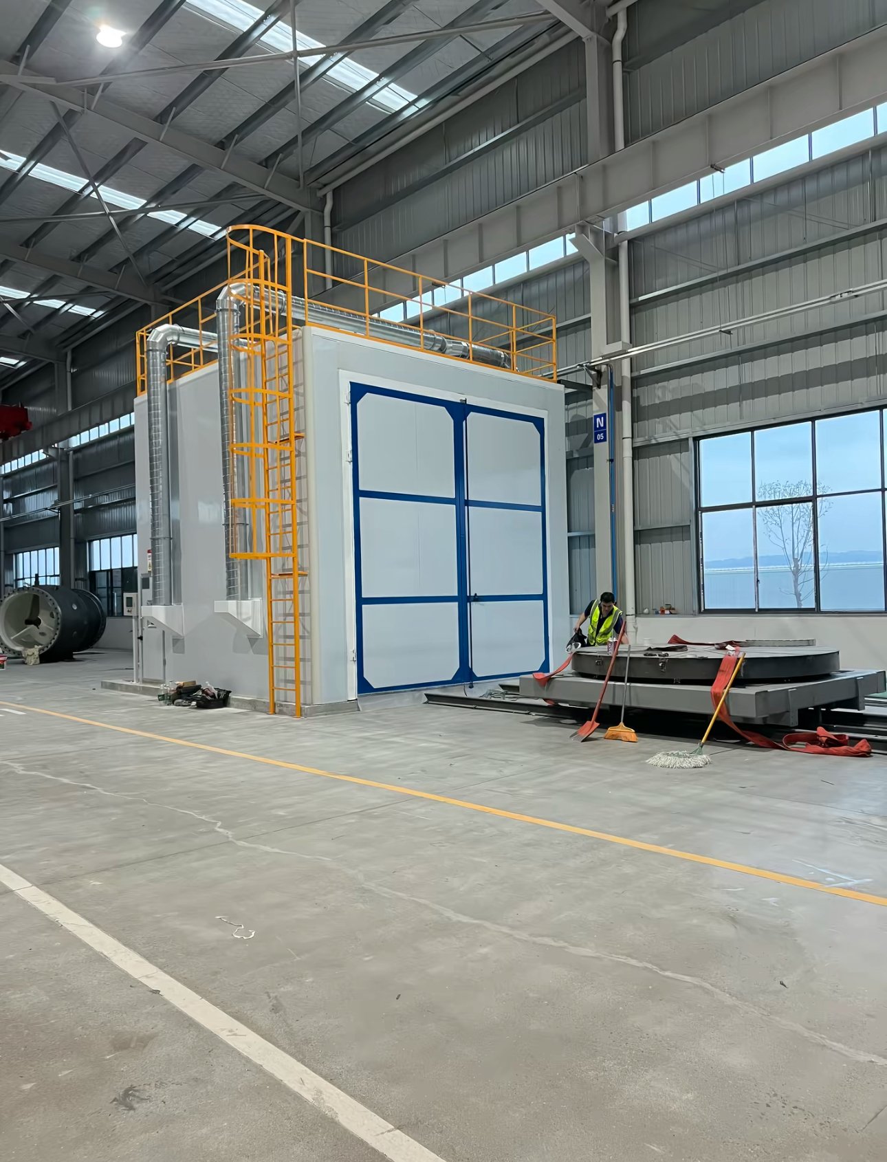 Sandblasting room with blue-framed double doors, exhaust ducts and a rail-mounted turntable for large workpieces in front
