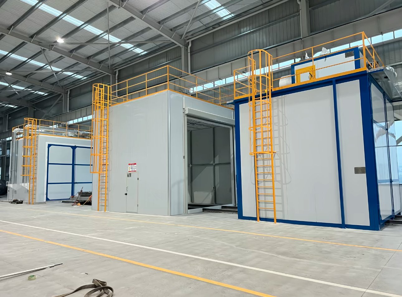 Three enclosed rooms of a blasting and painting line with roof platforms, yellow access ladders and blue-framed doors in a steel workshop