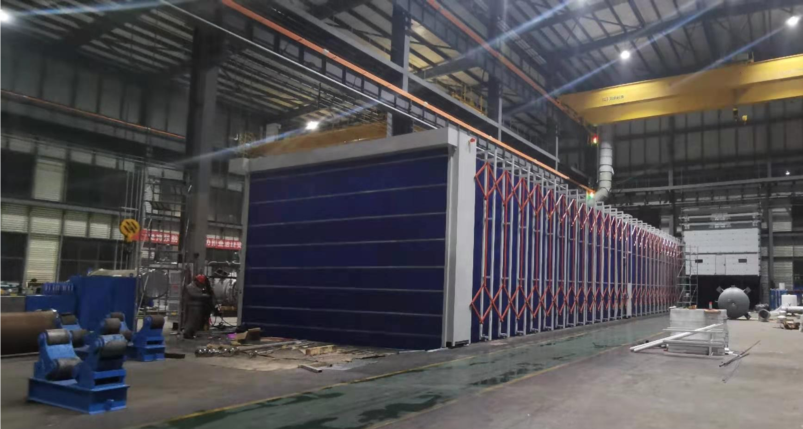 Long telescopic spray booth with blue flame-retardant PVC fabric and orange folding frames in a heavy fabrication hall with overhead cranes
