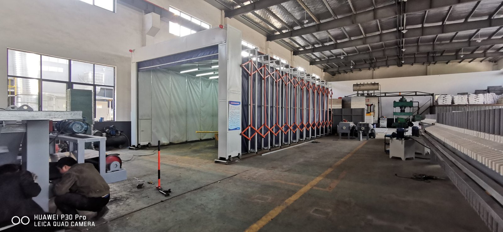 Side view of a telescopic spray booth with folded frames and fabric, control cabinet on the fixed end, in a machinery workshop
