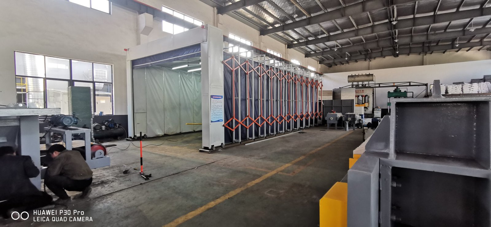 Telescopic spray booth with orange folding frames retracted in a machinery workshop, operators working beside the rail