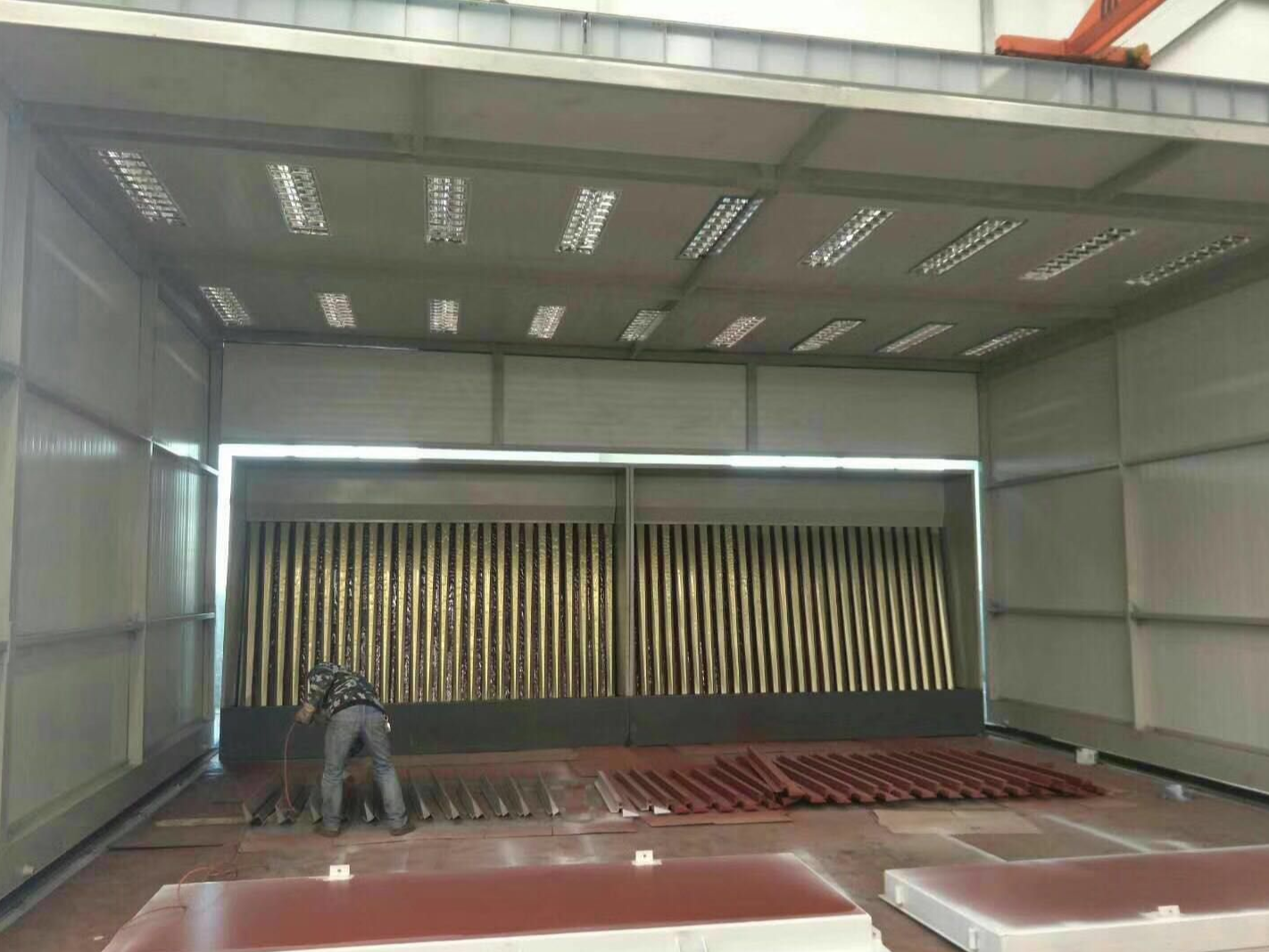 Interior of a large telescopic spray booth with corrugated dry filter wall, rows of ceiling lights and an operator preparing steel profiles