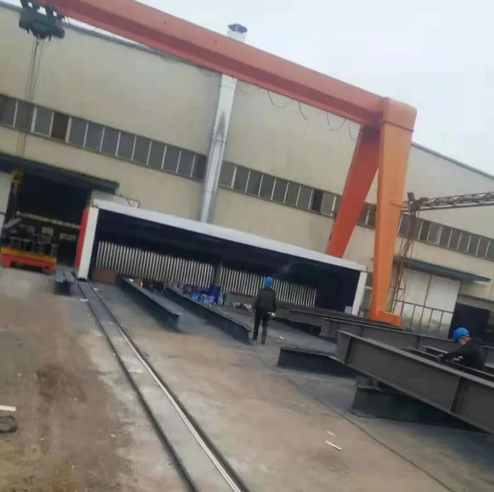 Cabine de pulvérisation télescopique sur rails au sol à l'extérieur d'un atelier sous un pont roulant, avec des structures en acier disposées pour la peinture